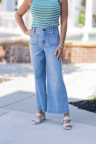 Mid-shot focusing on the lower body to showcase the front details of the Dear John Audrey Eclipse Jeans. The light-wash, high-rise denim features distinctive vintage-inspired twin open front patch pockets, a dark center top button, and a structured wide-leg cut. The pants cascade down into a cropped length with a tailored clean hem, styled with off-white heeled buckle sandals on a concrete patio walkway.