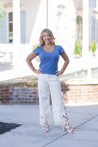 A full-length outdoor portrait of a smiling woman displaying the Dear John Audrey Lace Jeans. These premium cream-colored denim trousers feature a structured high-rise waist and a relaxed wide-leg silhouette. The true highlight is the intricate, bohemian-inspired cutout lace work along the lower leg that finishes in a dramatic scalloped hemline. She styles the statement jeans with a tucked-in slate blue ribbed t-shirt and cream block-heeled buckle sandals against a sunny courtyard backdrop.