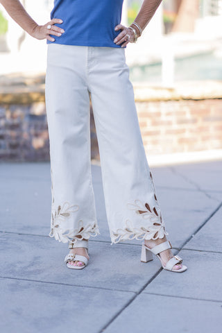 Mid-shot focusing on the legs to capture the detailed front craftsmanship of the Dear John Audrey Lace Jeans. The relaxed wide-leg cut is highlighted in a versatile cream colorway, accentuating the artistic open-work lace patterns sweeping across the lower shins. The un-distressed fabric ends beautifully at the contoured, feminine scalloped cuffs, paired with off-white block-heeled sandals.