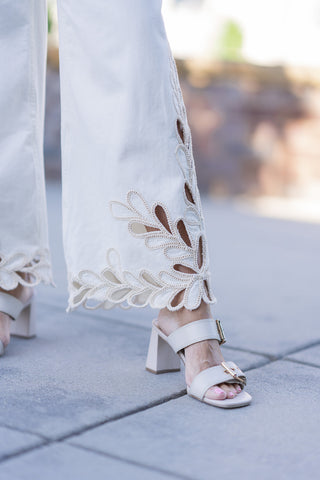 A detailed macro view focusing on the intricate cutout lace work and scalloped hemline of the Dear John Audrey Lace Jeans. The shot emphasizes the high-end boutique quality of the embroidered, leaf-like open lace motifs along the lower leg and the clean, secure stitching on the cream stretch denim fabric. A cream leather sandal with metallic buckle detailing is visible at the bottom of the frame.