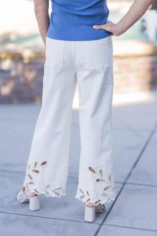 Back view of the Dear John Audrey Lace Jeans highlighting the smooth, flattering fit of the high-rise waistband and traditional functional back pockets. The high-quality stretch denim in a refined cream hue flows down to a wide leg, showcasing the stunning cutout leaf-patterned lace detailing and embroidered scalloped edges at the lower back hem. The model stands with one hand on her hip to display the relaxed yet polished silhouette.