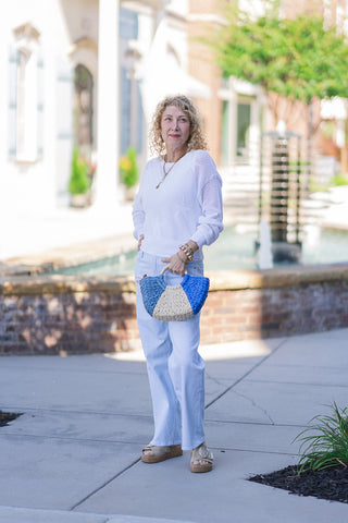 A full-length outdoor shot of a woman modeling the Dear John Skyway Taylor Jeans. The pants feature a crisp light blue and white vertical pinstripe pattern with a straight-to-wide-leg silhouette and an un-distressed clean hem. Styled for a coastal resort look, she pairs the denim trousers with a lightweight white open-knit star sweater, platform cork sandals, and a blue and neutral woven straw tote bag.