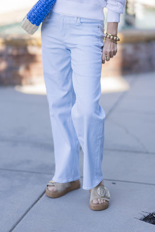 A cropped, close-up view focusing on the tailored details of the Dear John Skyway Taylor Jeans. The shot emphasizes the rich texture of the premium stretch denim and the fine light blue and white pinstripe pattern. It highlights the traditional five-pocket design at the hips and follows the relaxed, wide-leg cut down to the un-distressed hem, paired with tan cork-sole slides.