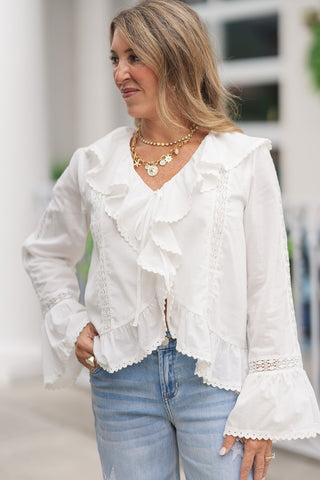 An upper-body portrait view of the Angelica Lace & Ruffle Blouse highlighting its feminine, whimsically detailed bodice. The off-white lightweight woven fabric drapes into a wide ruffled V-neckline with a delicate front tie closure. The sheer vertical lace inserts on the front panels and the lace-trimmed trumpet sleeves are prominently displayed. The woman poses with one hand in her pocket, accessorized with a layered gold charm necklace.