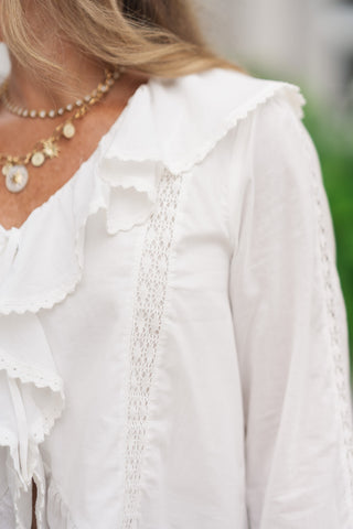 A detailed close-up shot focusing on the delicate fabrics of the Angelica Lace & Ruffle Blouse. The image highlights the intricate texture of the vertical lace insert panel running down the front bodice, contrasted against the crisp woven fabric. The soft, scalloped embroidery along the cascading ruffle trim is visible next to a gold link chain necklace, showcasing the high-end boutique craftsmanship.