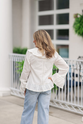 Back view from the waist up of a woman wearing the Angelica Lace & Ruffle Blouse, showcasing the romantic design elements from behind. The off-white blouse features a wide, ruffle-trimmed collar that drapes over the shoulders, a single vertical lace insert strip running down the center spine, and a gathered, ruffled flounce hem at the waist. The model poses with one hand on her hip, highlighting the flared cuffs and clean fit against light-wash blue jeans.
