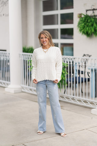 A full-length front view of a woman standing on an outdoor patio wearing the Dear John Rania sweater. The long-sleeve knit pullover is featured in a warm cream colorway, showcasing a slouchy, relaxed fit, a ribbed crew neckline, and a slightly cropped hemline with distressed fringed detailing. It is styled for an effortless Americana look with light-wash straight-leg denim jeans featuring star patch appliques, cream flat slide sandals, and a gold link charm necklace.