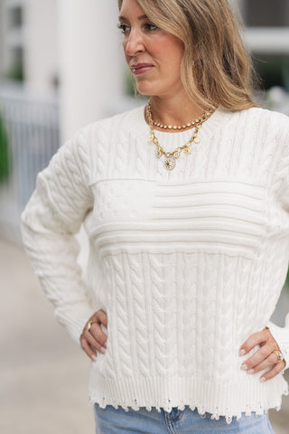 A portrait close-up shot focusing on the high-end detailing of the Dear John Rania sweater. The image highlights the structure of the integrated tonal American flag motif on the chest, surrounded by classic cable-knit stitching. The woman poses with her hands on her hips, showcasing the subtle fringe distressing along the ribbed crew neck and the relaxed drape of the dropped shoulders.