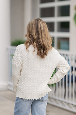 Back view from the waist up of a woman wearing the Dear John Rania sweater, emphasizing the cozy texture and fit from behind. The continuous cream cable-knit pattern runs seamlessly down the back panel and sleeves to a distressed, frayed ribbed hem. The pullover is styled loose to demonstrate its non-bulky drape over the back pockets of her high-waisted light-wash jeans.