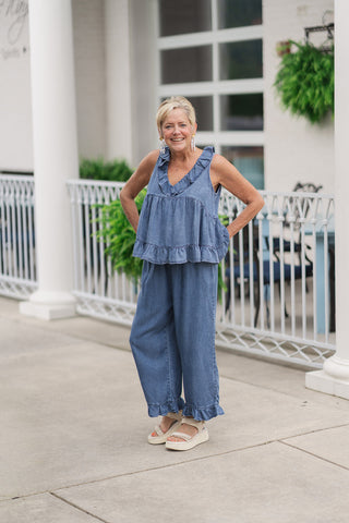 A smiling woman posing on an outdoor patio wearing the Southern Belle Babydoll Top, a lightweight sleeveless denim top in a vintage-inspired medium blue acid wash. The top features a tiered, flared babydoll silhouette with a dramatic ruffle trim framing the V-neckline. She has styled it as a coordinated set with the matching wide-leg Darlin' Denim Frill Capris, cream-colored platform wedge sandals, and statement pearl drop earrings.