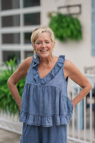 An upper-body portrait view of the Southern Belle Babydoll Top highlighting its romantic details. The soft, breathable tencel-blend denim fabric is shown in a textured medium wash, emphasizing the prominent frill trim that borders the flattering plunging V-neckline. The tiered bodice creates a flirty, flared shape that drapes beautifully, accessorized with gold flower studs and cascading pearl drop earrings.