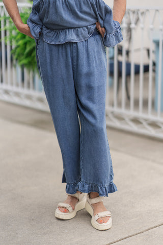 A mid-shot view from the waist down focusing on the front details of the Darlin' Denim Frill Capris. The image highlights the relaxed, wide-leg cropped silhouette that drapes fluidly down to a playful ruffled frill hemline. The comfortable, flat-front elasticized waistband sits smoothly under the ruffled edge of the matching denim top, styled with cream open-toe platform slide sandals.