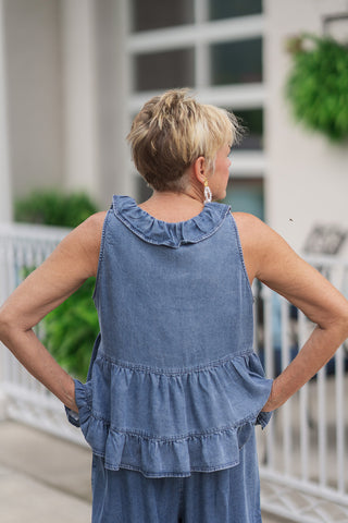 Back view from the waist up of a woman wearing the Southern Belle Babydoll Top, showcasing the continuous details from behind. The statement ruffle collar wraps elegantly around the back of the neck, while the relaxed, tiered bodice flows into a playful ruffled bottom hemline. The model poses with her hands on her hips, demonstrating the loose, comfortable, and non-clinging fit of the lightweight chambray top.