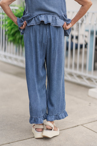 Back view from the waist down of a woman wearing the Darlin' Denim Frill Capris, showcasing the fit and elasticized gathering of the waistband from behind. The vintage-inspired medium blue denim fabric drapes loose through the legs, highlighting the continuous ruffled frill hem at the cropped cuffs. The model poses with her hands on her hips, showing the back-elastic details of the pull-on style.