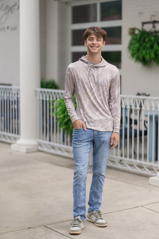 A full-length front view of a male model smiling while posing in the Performance Camo Drift Hoodie. The lightweight men's activewear hoodie features a straight hemline that drapes cleanly over light-wash, slim-fit denim jeans. He completes the casual lifestyle look with neutral green and beige suede sneakers, standing on an outdoor sidewalk in front of a white building fence with green hanging plants.