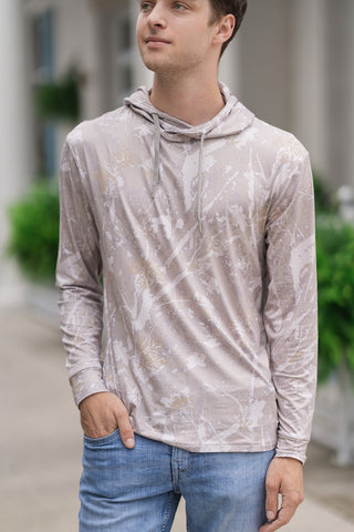 A close-up view highlighting the detailed features of the Performance Camo Drift Hoodie on a male model. The image showcases the unique abstract drifter camo pattern in a muted color palette of tan, soft grey, and off-white. The modern crossover hooded neckline is visible, complete with matching light grey drawstrings and a smooth, ultra-lightweight performance fabric construction. The model has one hand tucked into the pocket of light-wash blue jeans against a softly blurred outdoor patio background.
