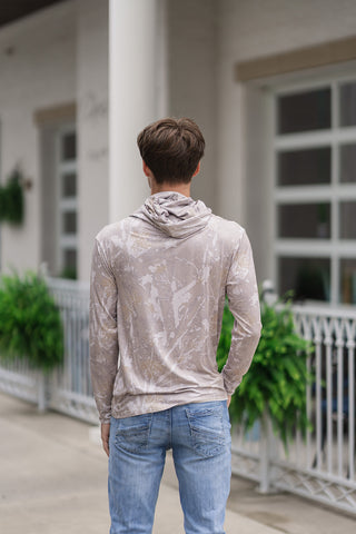 A full-length back view of the Performance Camo Drift Hoodie, showcasing its clean-cut tailored drape and flexible fit. The image highlights the continuous nature-inspired abstract camo print across the shoulders and long sleeves, alongside the relaxed structure of the crossover hood resting on the upper back. The straight bottom hem sits neatly over light-wash blue jeans as the model stands outdoors looking toward a building entrance.
