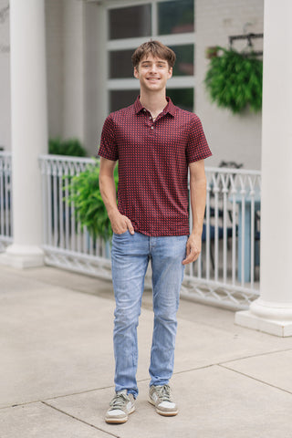 A full-length front view of a male model smiling while presenting the Georgia Performance Polo. The short-sleeve polo features a vibrant allover geometric pattern of red Georgia state silhouettes against a dark maroon background. It is styled untucked with a straight hemline draped over light-wash, slim-fit denim jeans. The casual summer lifestyle look is completed with neutral-toned green and beige fashion sneakers on an outdoor sidewalk with a white building facade and fence in the background.