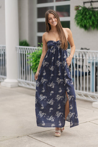 Model wearing the Midnight Garden Strapless Maxi Dress, a floor-length strapless empire waist dress featuring a dark navy textured fabric printed with muted cream floral botanical graphics. Styled with platform woven espadrille heels outdoors, demonstrating the flowy silhouette, subtle front leg slit, and functional side hand pocket.