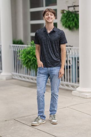 A full-length front view of a male model smiling while presenting the Black Camo Performance Polo. The short-sleeve polo features a straight hemline that fits true to size, styled untucked over light-wash, slim-fit denim jeans. He completes the versatile lifestyle look with neutral green and beige fashion sneakers, standing outdoors on a sidewalk next to a white structural column and a white fence decorated with green plants.