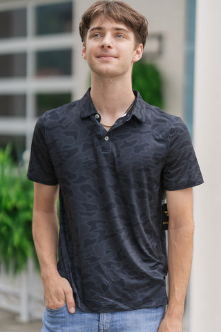 A portrait-style close-up highlighting the structural details of the Black Camo Performance Polo on a male model. The image showcases the subtle, tone-on-tone dark camo pattern in shades of black and dark grey. The classic detailing is on display, featuring a crisp structured collar and a clean three-button front placket, slightly unbuttoned to reveal a gold chain necklace. The lightweight performance material fits smoothly over his shoulders as he poses with one hand in his pocket against a blurred patio.
