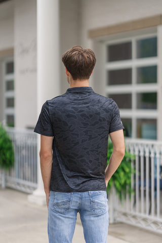 A full-length back view of the Black Camo Performance Polo, highlighting its polished fit and modern athletic silhouette. The image shows the continuous tonal black and grey camouflage print running seamlessly across the back panel and short sleeves. A small, subtle brand logo is embroidered just below the rear collar seam. The straight bottom hemline sits naturally over light-wash blue jeans as the model stands facing away toward a building entrance.