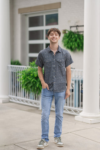 A full-length front view of a male model smiling while presenting the Paisley Spurs Performance Western Shirt. The short-sleeve button-down shirt is styled untucked, displaying its tailored drape over light-wash, slim-fit denim jeans. The casual summer outfit is rounded out with neutral green and beige fashion sneakers on a concrete patio walkway flanked by white pillars and a building fence decorated with greenery.