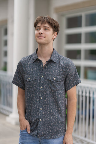 A portrait-style mid-shot highlighting the front tailoring of the Paisley Spurs Performance Western Shirt on a male model. The performance shirt features a detailed allover grey and cream paisley pattern layered with western motifs against a slate-charcoal backdrop. The design showcases a clean-cut standard collar, a full front placket with secure closures, and two structured front patch pockets with snap-button flaps. 