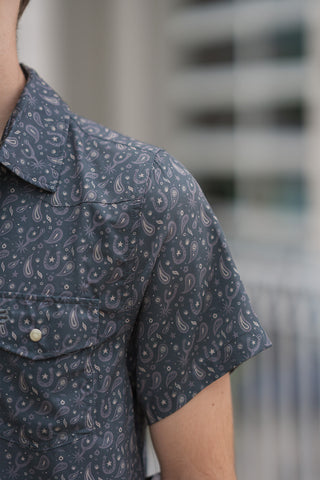 A close-up macro shot showcasing the intricate graphic print and premium stitching of the Paisley Spurs Performance Western Shirt. The slate-charcoal fabric is finely detailed with micro cream and grey traditional western motifs, including horseshoes, riding spurs, cowboy hats, and desert stars woven into a grid-like paisley layout. The image clearly displays a dual chest pocket flap adorned with a secure pearlized snap button, alongside the pointed stitching of the western front shoulder yoke.