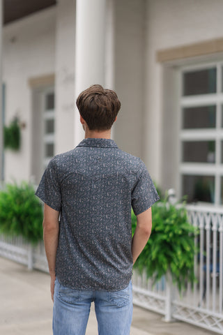 A full-length back view of the Paisley Spurs Performance Western Shirt, emphasizing its authentic cowboy silhouette and broadening shoulder lines. The continuous grey and cream paisley print runs seamlessly across the back panel and short sleeves, highlighted by a traditional pointed western back yoke stitching detail. The straight bottom hemline falls cleanly over light-wash blue jeans as the model stands facing away toward a modern building entrance.
