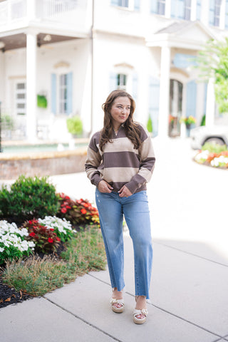 A full-length view of a woman posing outdoors wearing the Iced Mocha Bold Stripe Knit—a sporty, preppy half-zip sweater styled with bold, wide horizontal stripes in rich espresso brown and creamy oat beige. Designed with a relaxed, boxy fit and low drop shoulders, the knit pullover is paired with light-wash straight-leg jeans with raw hems and off-white platform espadrille sandals.
