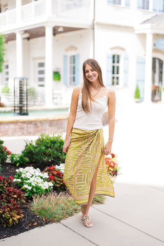 Woman smiling in the Laura Wrap Skirt, an asymmetrical faux wrap midi skirt featuring a graphic abstract print in mossy green, chartreuse, and blush pink. Styled for summer with a simple white spaghetti strap tank top, off-white flat thong sandals, and a delicate gold chain necklace while standing on a paved walkway near lush flowerbeds and Mediterranean-style resort architecture.