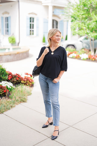 A full-length view of a woman wearing the Black Cloud V-Neck Top, an ultra-soft, lightweight black sweater tee featuring wide elbow-length sleeves and a flattering V-neckline. Styled effortlessly for a casual chic look with light-wash straight-leg jeans, black pointed-toe slingback heels, a chunky gold coin pendant necklace, and a black shoulder bag, set against an elegant outdoor shopping courtyard backdrop.