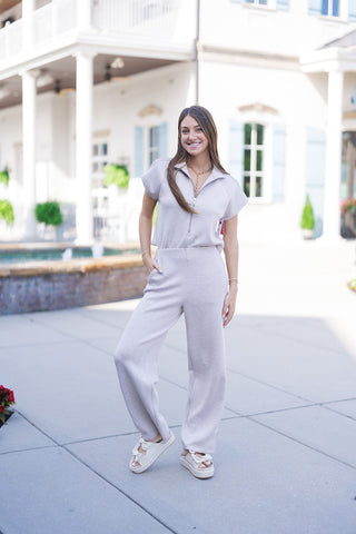 Woman smiling in the SPANX Pintuck Half Zip Jumpsuit, a short-sleeve activewear jumpsuit in a soft oatmeal heather beige tone. The one-piece suit features a silver half-zip front, high open collar, pintuck shoulder details, cap sleeves, and relaxed straight legs with side pockets. Styled for casual athleisure with off-white platform slide sandals and a gold chain necklace outdoors.
