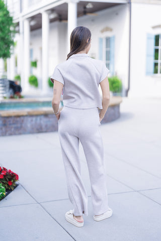Full-length back view of the SPANX Pintuck Half Zip Jumpsuit in oatmeal heather beige, showing the relaxed fit across the back, extended cap sleeves with raglan seams, and straight wide-leg drape down to the ankles as the model stands turned away with hands in her pockets.
