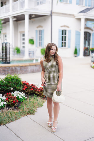 Woman posing in the Halle Mini Dress, a retro-inspired sleeveless mini dress in a rich olive green shade adorned with a crisp cream polka dot pattern. The tailored shift silhouette features a modest high boat neckline and subtle contouring seams along the bodice. Styled for an elevated daytime look with off-white platform espadrille wedge sandals, a textured cream lace handbag, a gold link necklace with a pearl drop, and pearl drop earrings near a coastal resort building.