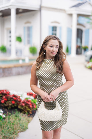 A portrait view of the Halle Mini Dress highlighting the structured high boat neckline and delicate bust dart details. The cream polka dots pop against the olive green fabric, complemented by a gold chain necklace with a freshwater pearl pendant, matching pearl drop earrings, and soft brown waves. The model holds a curved cream lace handbag at her waist.