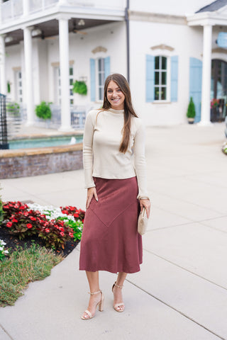 A smiling woman posing outdoors in the Champagne Sparkle Mock Neck Top—a warm cream champagne fitted long-sleeve knit top with fine vertical ribbing and subtle metallic shimmer threading throughout. Styled for a chic autumn look paired with a high-waisted burgundy faux-suede midi skirt, nude ankle-strap block heels, and a beige leather handbag in front of a white brick building.