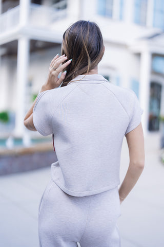 Upper body back angle shot showcasing the high shawl-style back collar, raglan shoulder construction, and subtle pintuck lines along the cap sleeves. The model is turned away touching her hair, highlighting the clean, unstructured back silhouette of the heather beige activewear top.