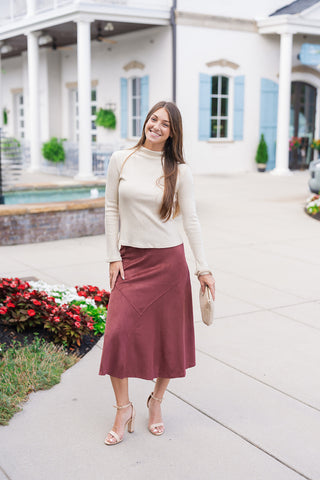 Woman smiling in the Espresso Elegance Suede Skirt, a high-waisted faux suede midi skirt in a rich mahogany brown hue. The skirt features a subtle mermaid silhouette that flares at the hem, detailed with unique diagonal asymmetrical seam paneling across the front. Styled for a sophisticated fall look with a long-sleeve cream ribbed mockneck top, nude ankle-strap block heel sandals, and a tan suede clutch bag in an outdoor plaza.