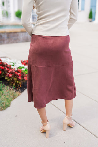 Back view of the Espresso Elegance Suede Skirt showcasing the smooth, high-waisted fit around the hips and a clean horizontal back seam running above the flared midi hemline. The model is turned away, wearing a fitted off-white long-sleeve ribbed top over the waistband and nude heel sandals.