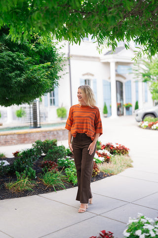Woman smiling while walking outdoors in the Pumpkin Spice & Everything Nice Sweater, a lightweight pumpkin-orange knit top with thin dark horizontal stripes. Designed with drop-shoulder elbow-length sleeves and a ribbed crew neckline, the relaxed pullover is front-tucked into high-waisted dark brown pants with front vertical seam details. Styled with neutral braided block-heel sandals under lush green tree branches in front of a resort backdrop.