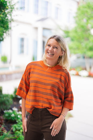 A close-up portrait of the Pumpkin Spice & Everything Nice Sweater showcasing the soft terracotta orange knit texture and subtle multi-line horizontal dark stripes. The shot highlights the thick ribbed crew neck collar, dropped shoulders, relaxed elbow sleeves with wide ribbed cuffs, and the gentle front tuck into high-waisted brown denim pants.