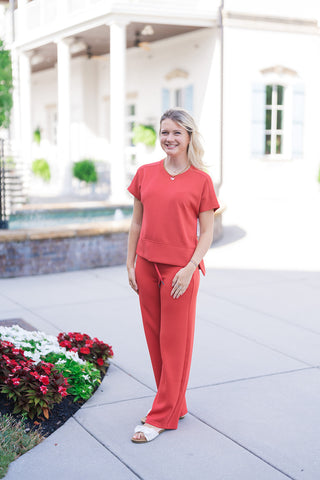 Woman smiling in the vibrant red SPANX Crewneck Tee, paired with matching red wide-leg drawstring pants for a coordinated lounge set. The short-sleeve top features a ribbed crew neckline, dropped shoulder seams, and a wide banded hem with subtle side slits. Styled with off-white flat bow sandals and layered gold necklaces outdoors in front of a white architectural backdrop.