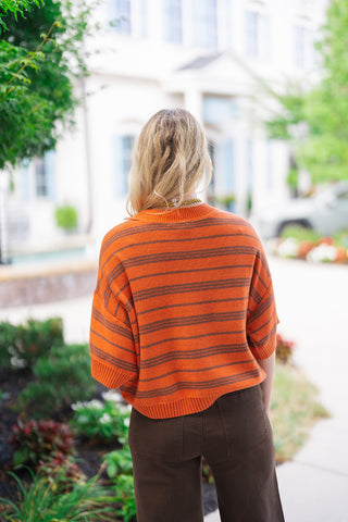 Back view of the Pumpkin Spice & Everything Nice Sweater displaying the boxy, relaxed knit silhouette and continuous dark horizontal stripes across the pumpkin orange fabric. The shot highlights the drop-shoulder construction, elbow-length sleeves with wide ribbed cuffs, and a ribbed straight hemline paired with high-waisted brown pants.