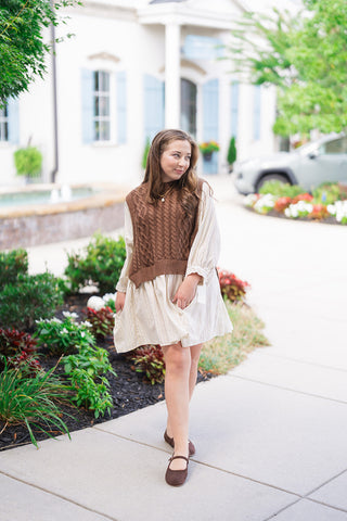 A woman posing outdoors in the Chilly Mornings Mini Dress, a layered two-in-one outfit featuring a cream and brown pinstriped A-line mini dress under a cropped chocolate brown cable-knit sweater vest. Styled for a preppy autumn look with dark brown ballet flats and a gold chain necklace in front of a boutique storefront.