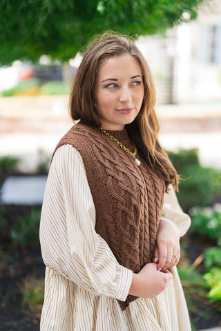 A portrait close-up view highlighting the detailed textures of the Chilly Mornings Mini Dress. The shot showcases the rich braided stitching of the chocolate brown cable-knit vest, the subtle olive and brown pinstripes on the cream base fabric, and the billowy 3/4-length balloon sleeves with gathered cuffs, accented by a gold pearl pendant necklace.