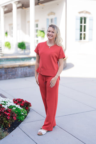 Woman posing in the SPANX Straight Leg Pants in a vibrant red hue, paired with a matching short-sleeve crewneck top for a bold monochromatic lounge set. The straight-leg trousers feature a comfortable pull-on design with an adjustable drawstring waist, paired with off-white flat bow sandals outdoors.