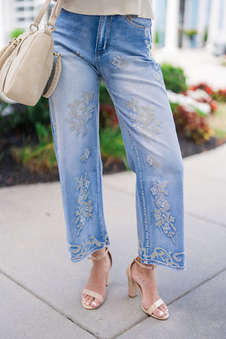 A mid-section close-up showcasing the front of the high-waisted Desert Rose Border Jeans. The shot details the medium blue denim wash, zip-fly waistband, and the cascading ivory floral embroidery motifs running down the front of both straight, relaxed legs to the ornate scrollwork embroidery border along the cropped ankle hemline.