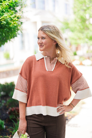 A mid-length portrait shot highlighting the front construction of the Spiced Chai Collar Knit Top. The view details the open V-neck placket lined with terracotta and cream stripes, the structured cream polo collar, the drop-shoulder seam detailing, and the wide cream ribbed hemline resting over high-rise brown pants.